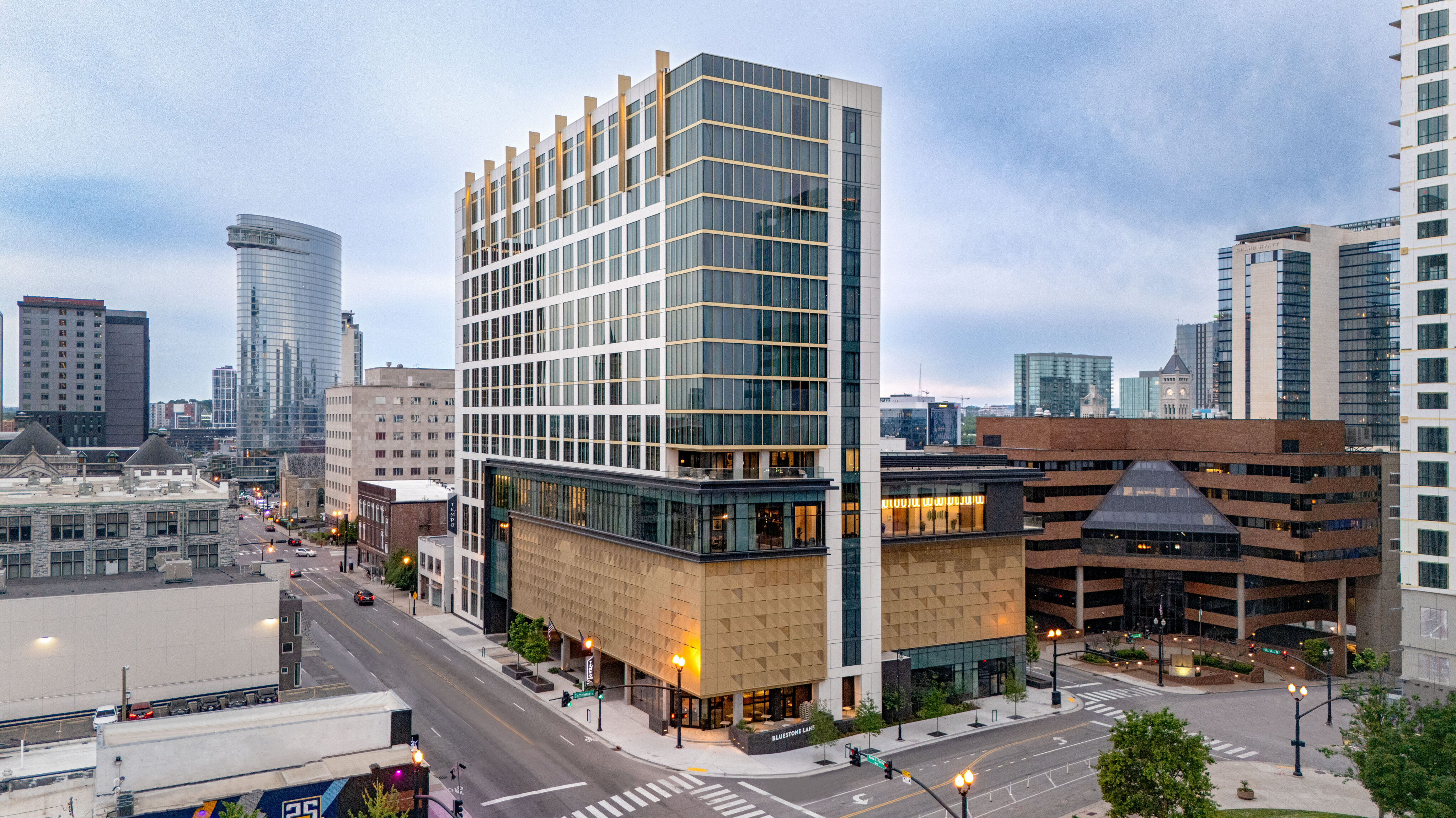 Modern mid-rise building with glass and tan exterior on a city corner, surrounded by other tall buildings and empty streets under a cloudy sky.