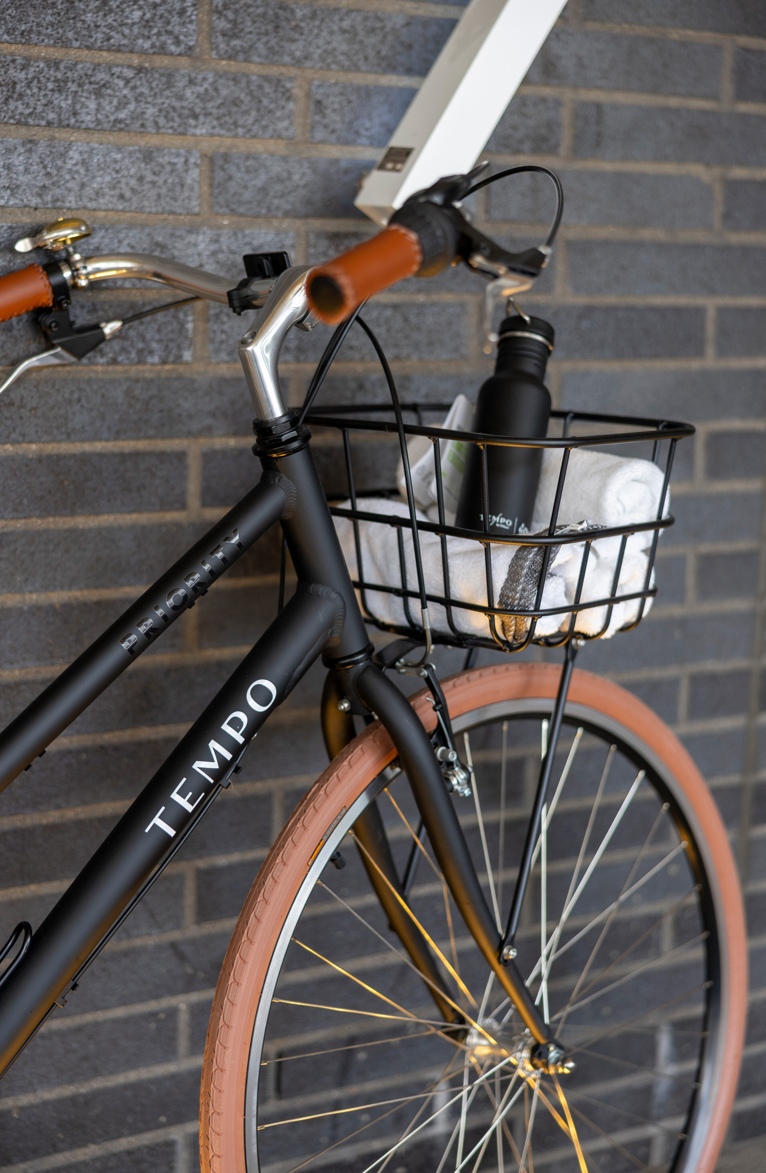 A black bicycle with tan tires is parked against a gray brick wall. A metal basket on the front holds a black water bottle and a white cloth.