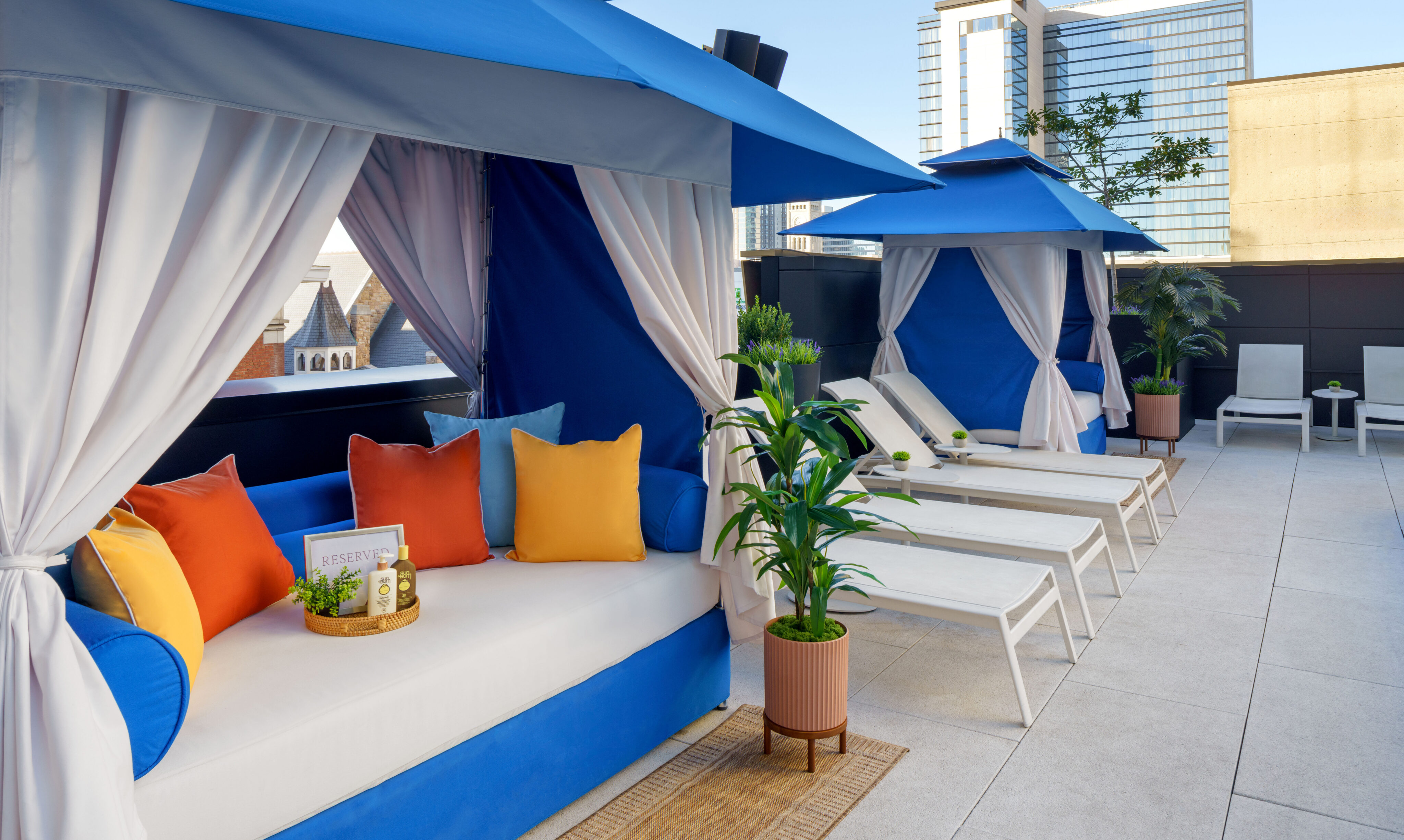 Rooftop lounge area with blue cabanas, white cushioned seating, colorful pillows, potted plants, and white lounge chairs beside a cityscape background.