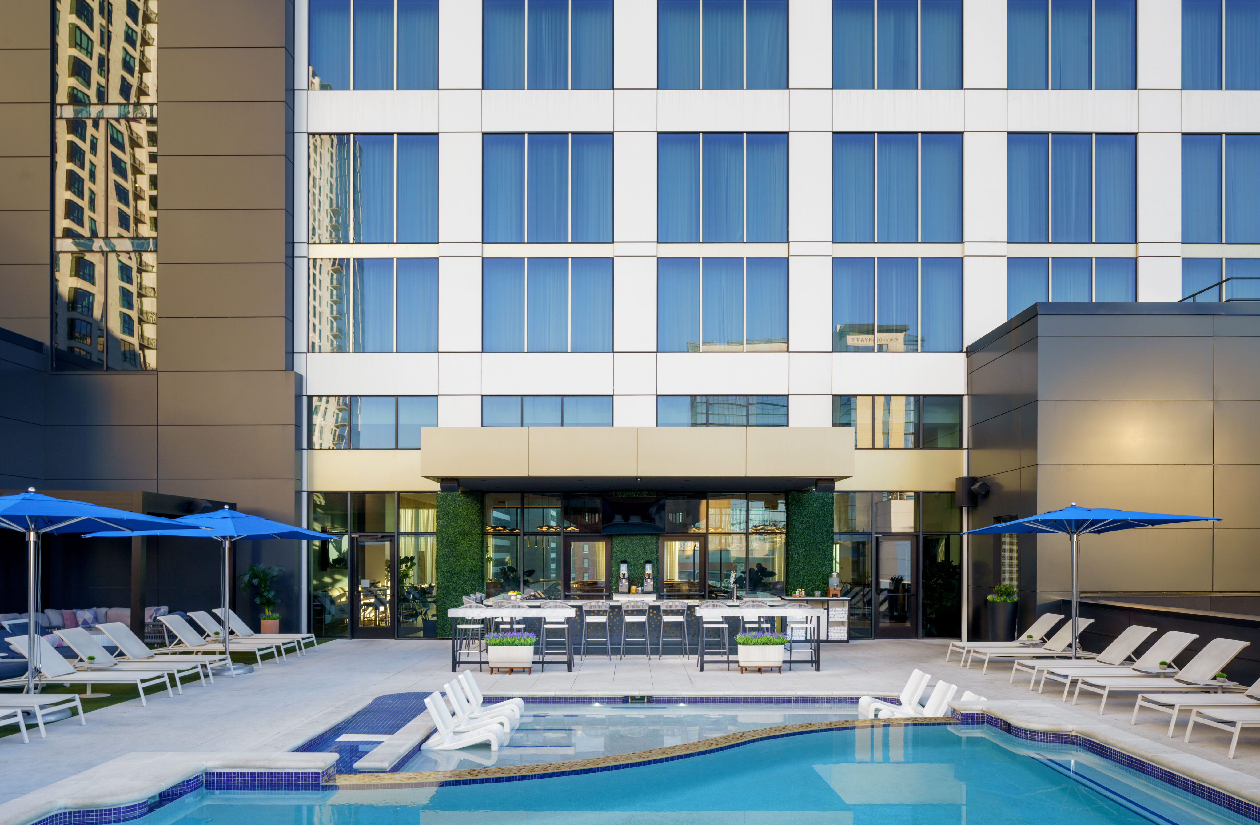Outdoor pool area with lounge chairs, tables, and blue umbrellas, set against a modern multi-story building with large windows.
