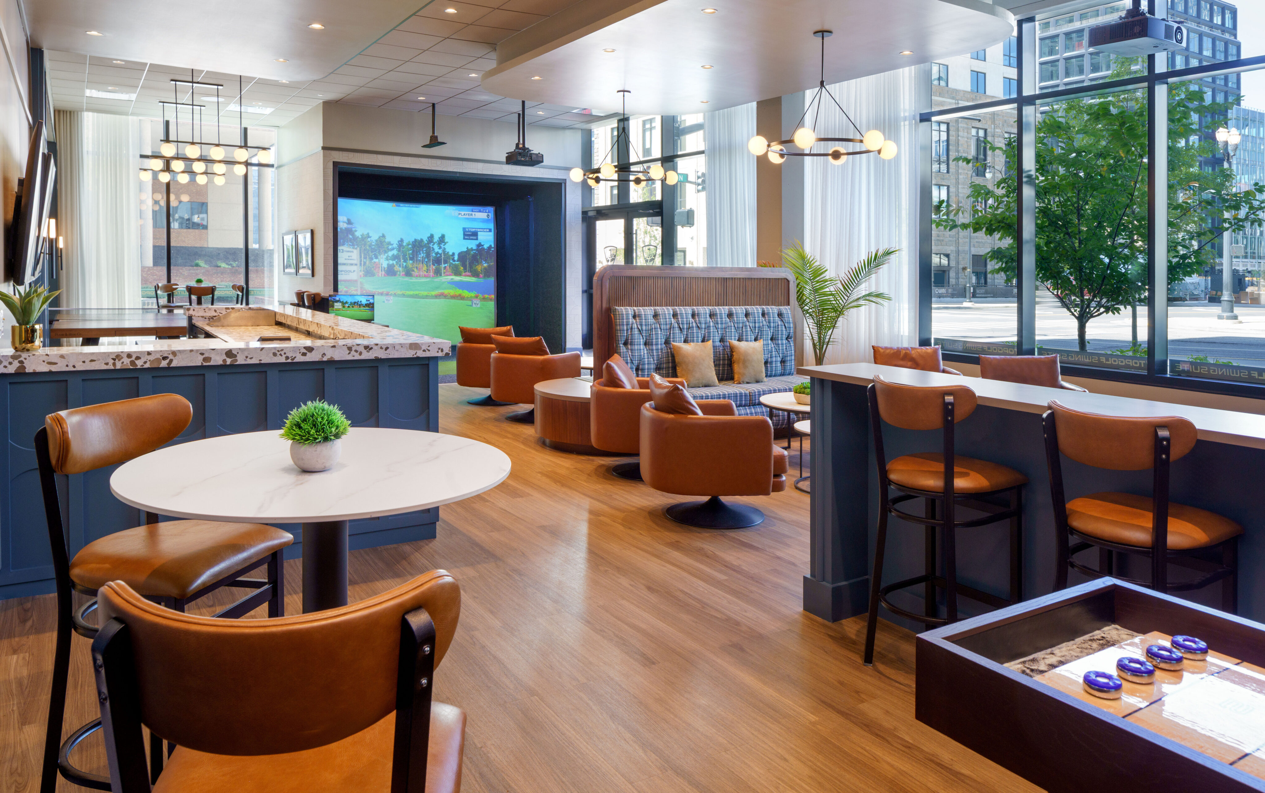 Modern lounge area with leather chairs, marble-topped tables, a bar counter, a tabletop game, and large windows overlooking city buildings and trees.