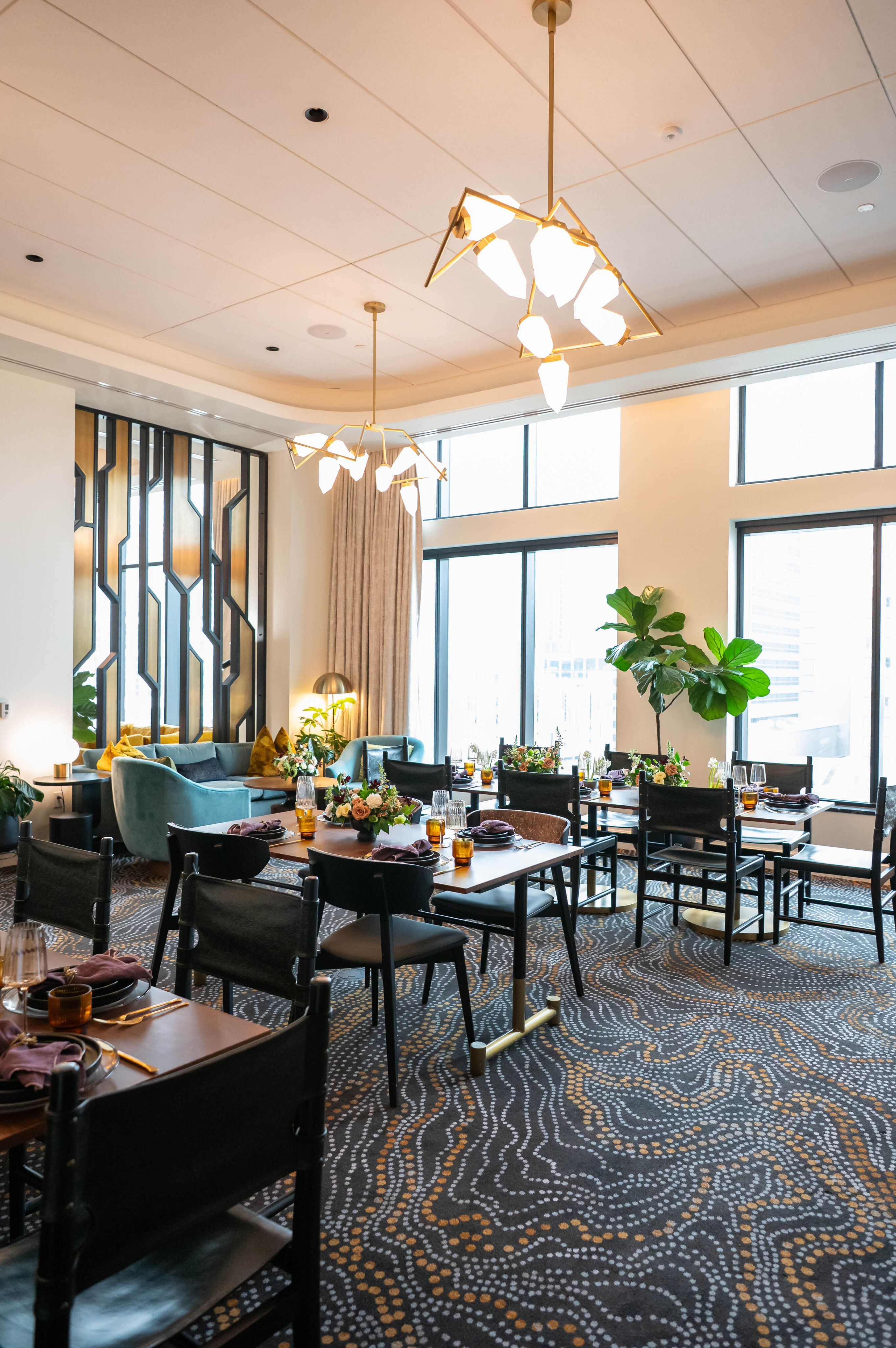 Modern dining room with patterned carpet, dark wood tables and chairs, decorative lighting, green plants, and large windows letting in natural light.