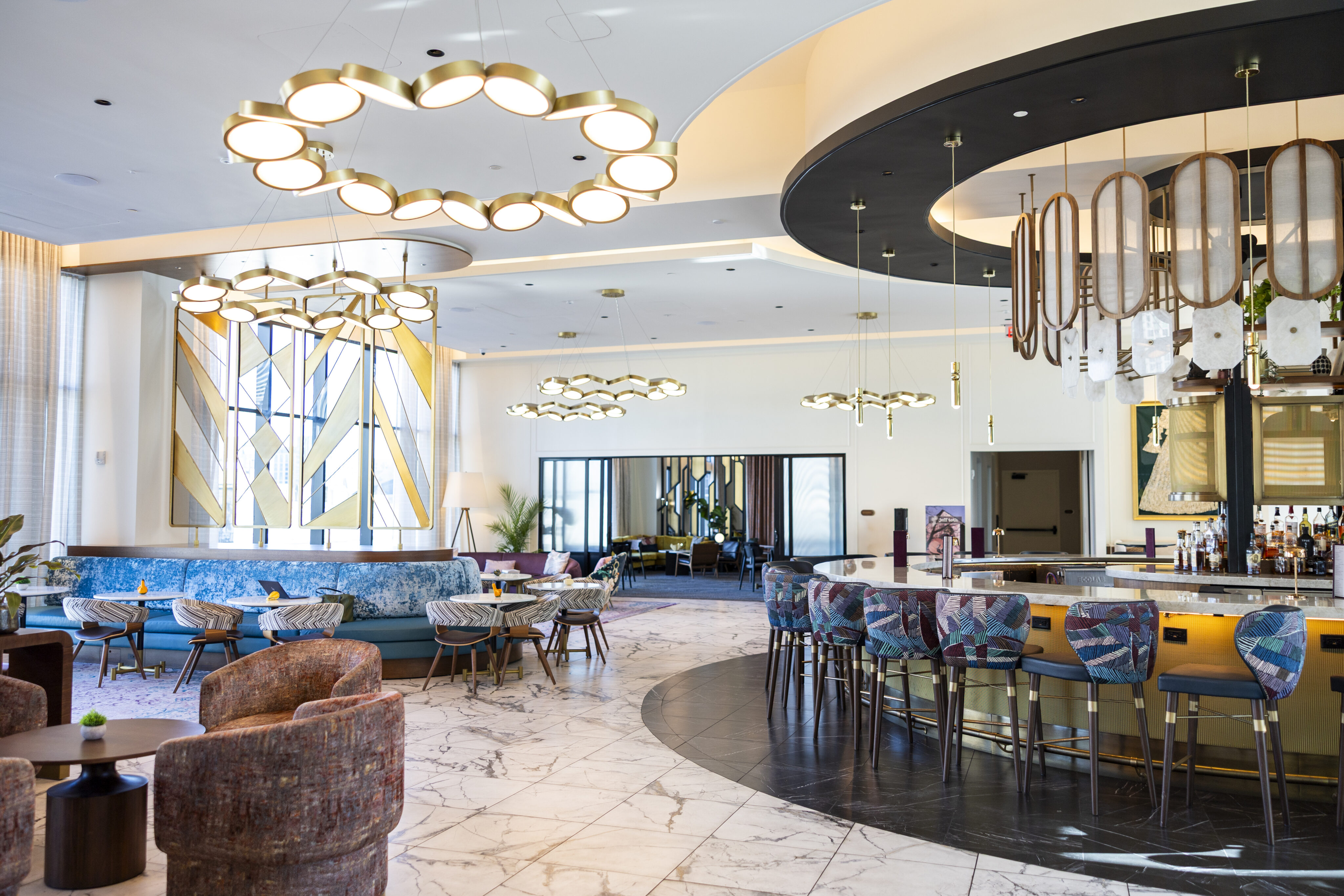 Modern hotel lounge with marble floors, curved bar with colorful stools, round chandeliers, patterned seating, and large geometric windows.
