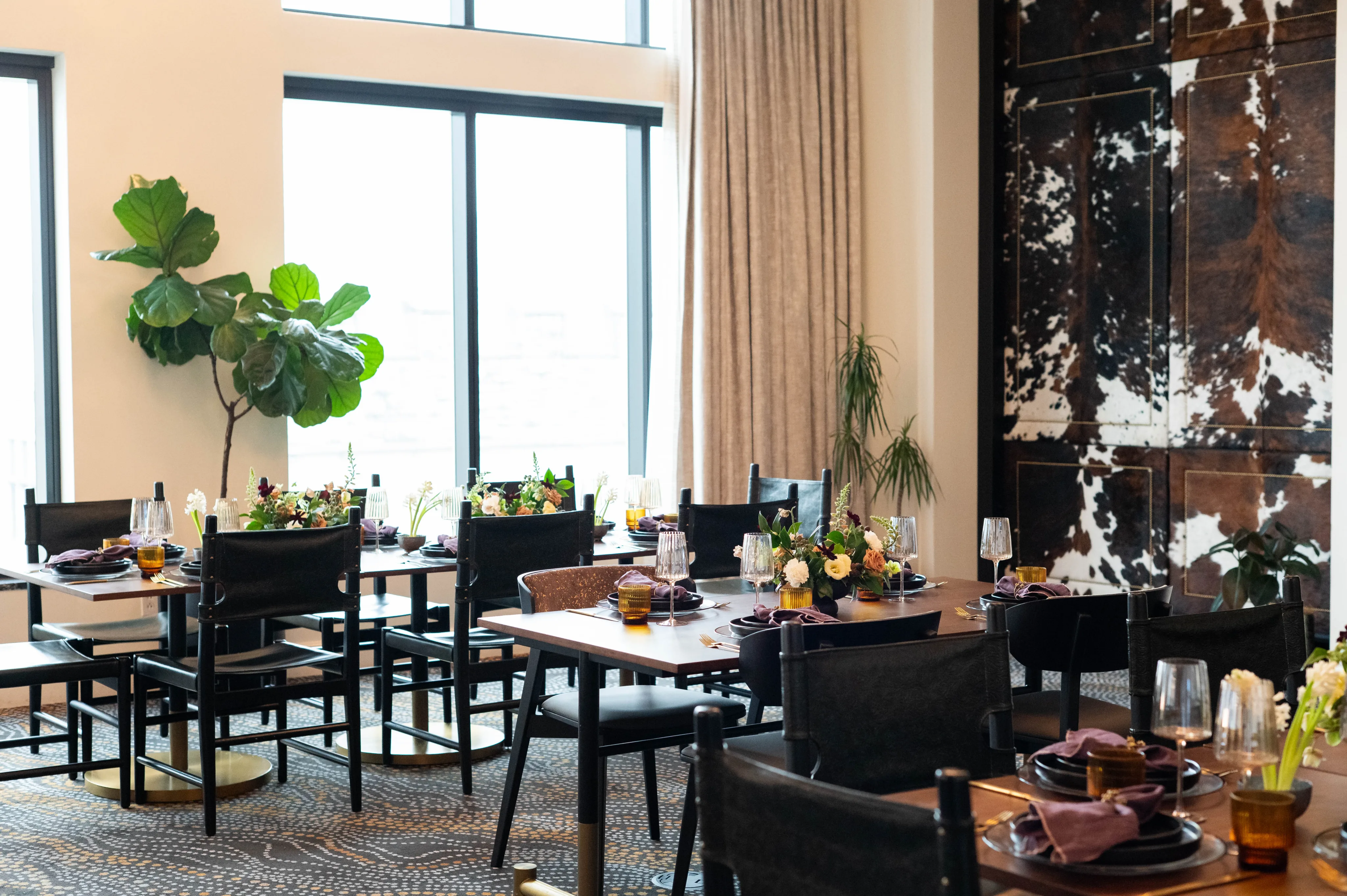A modern dining room with set tables, black chairs, floral centerpieces, and large windows with natural light. Perfect for Private Events, the space features plants near the windows and a decorative wall panel.