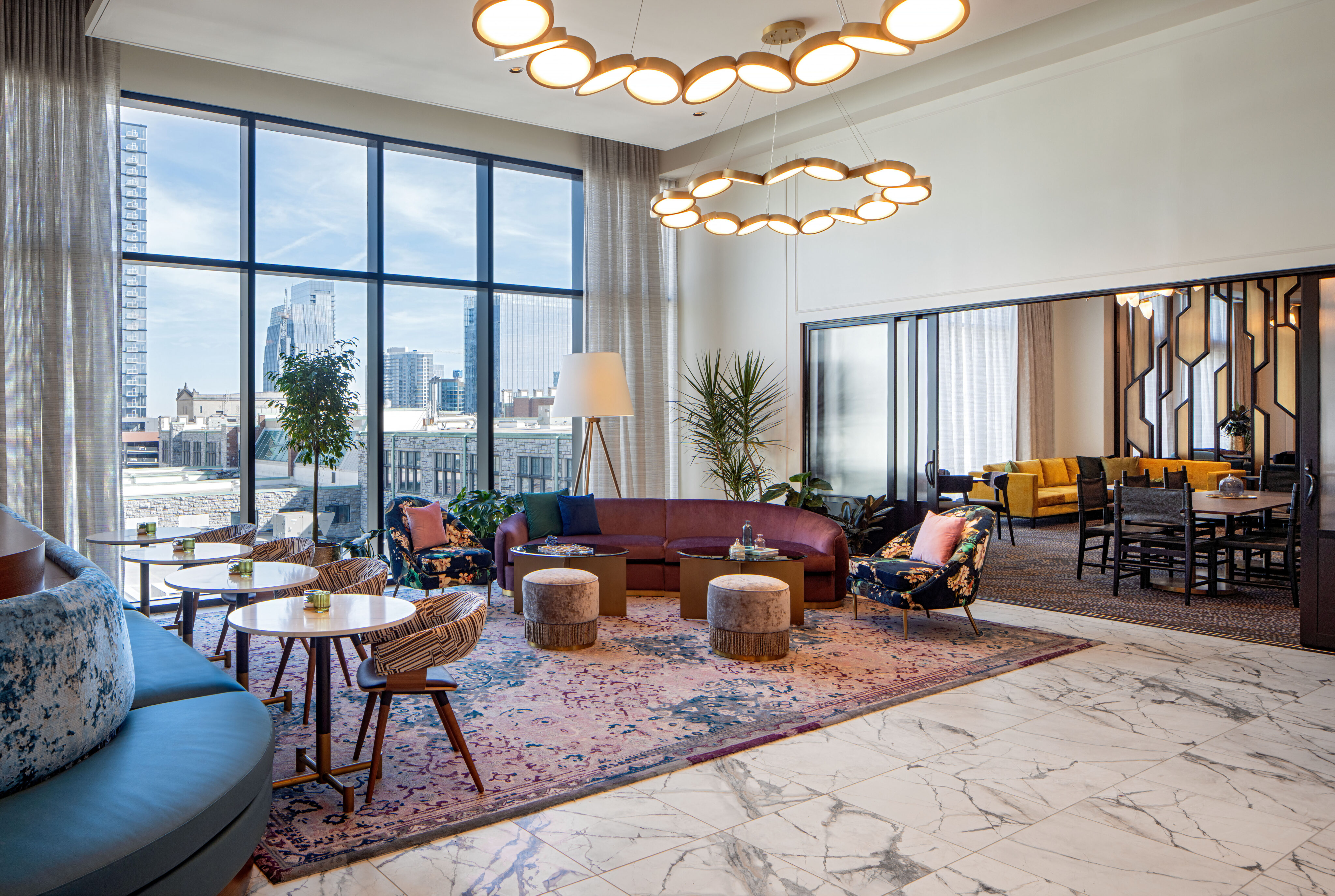 Lovelorn Lounge: Modern lounge area with large windows, city view, marble floor, colorful rugs, varied seating, round tables, and ceiling lights; adjacent room with dining table visible through a glass partition.