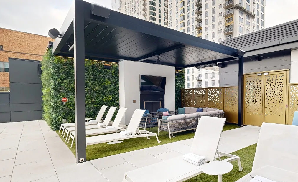 Rooftop lounge area with white lounge chairs, a covered seating area under a pool pergola, TV, green wall, and gold perforated doors, set against the backdrop of city high-rise buildings.