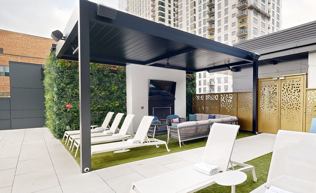 Rooftop lounge area with white lounge chairs, a covered seating area under a pool pergola, TV, green wall, and gold perforated doors, set against the backdrop of city high-rise buildings.