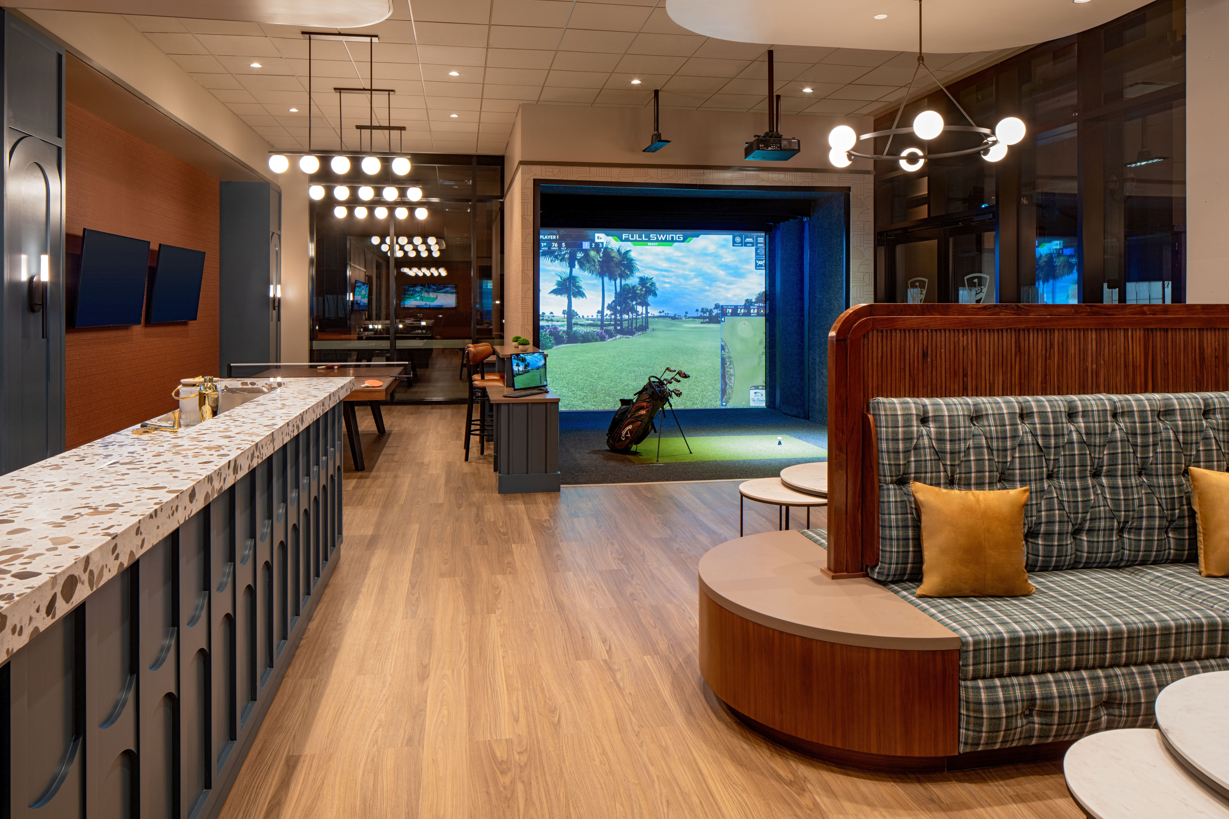 Modern lounge featuring a Topgolf Swing Suite with a golf simulator, bar counter, booth seating, and multiple TVs; golf clubs are set near the simulator screen displaying a course.