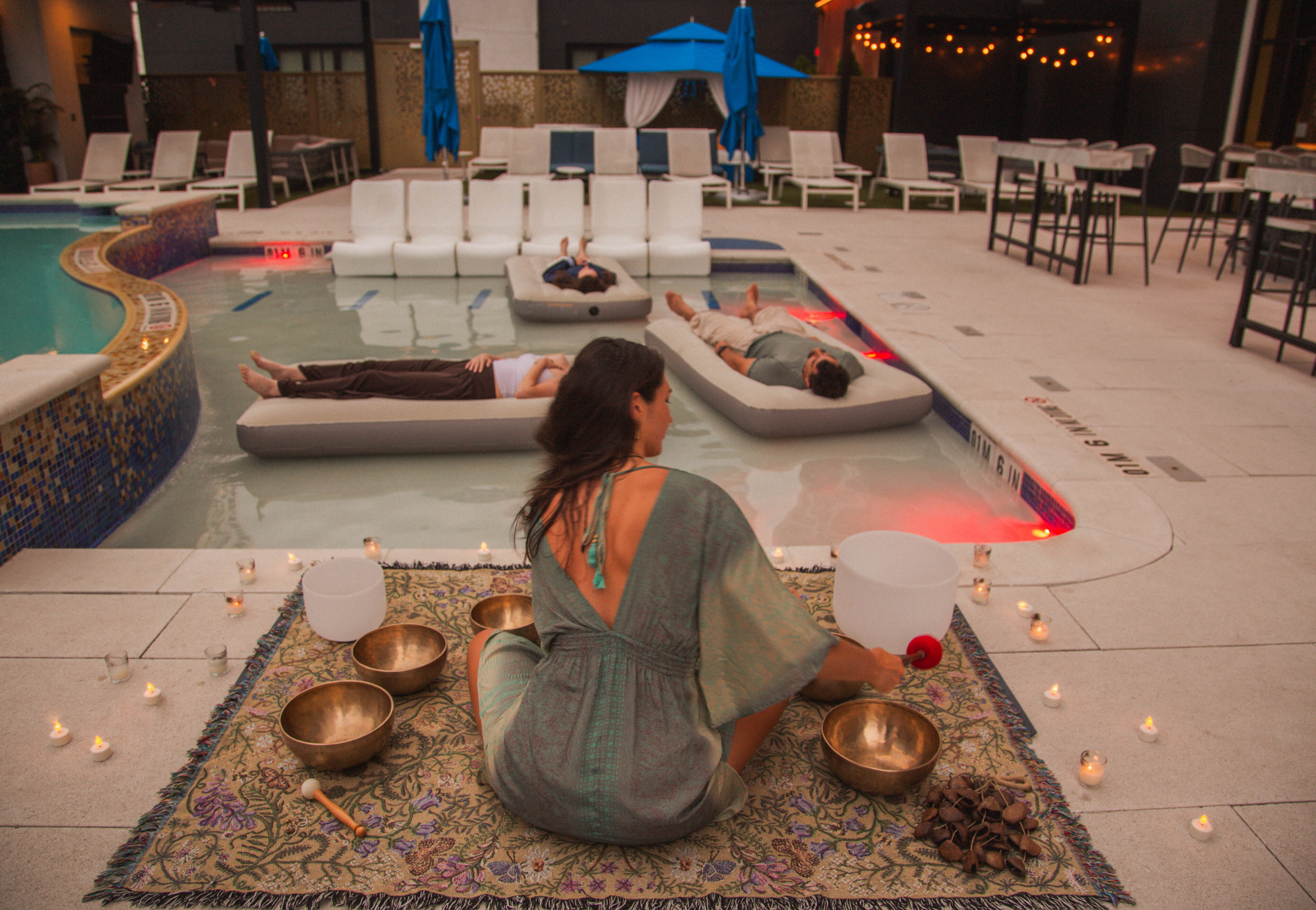 A woman leads a poolside soundbath, surrounded by singing bowls and candles, as three people relax on floating mats in the water, soaking in the calming floating frequencies.