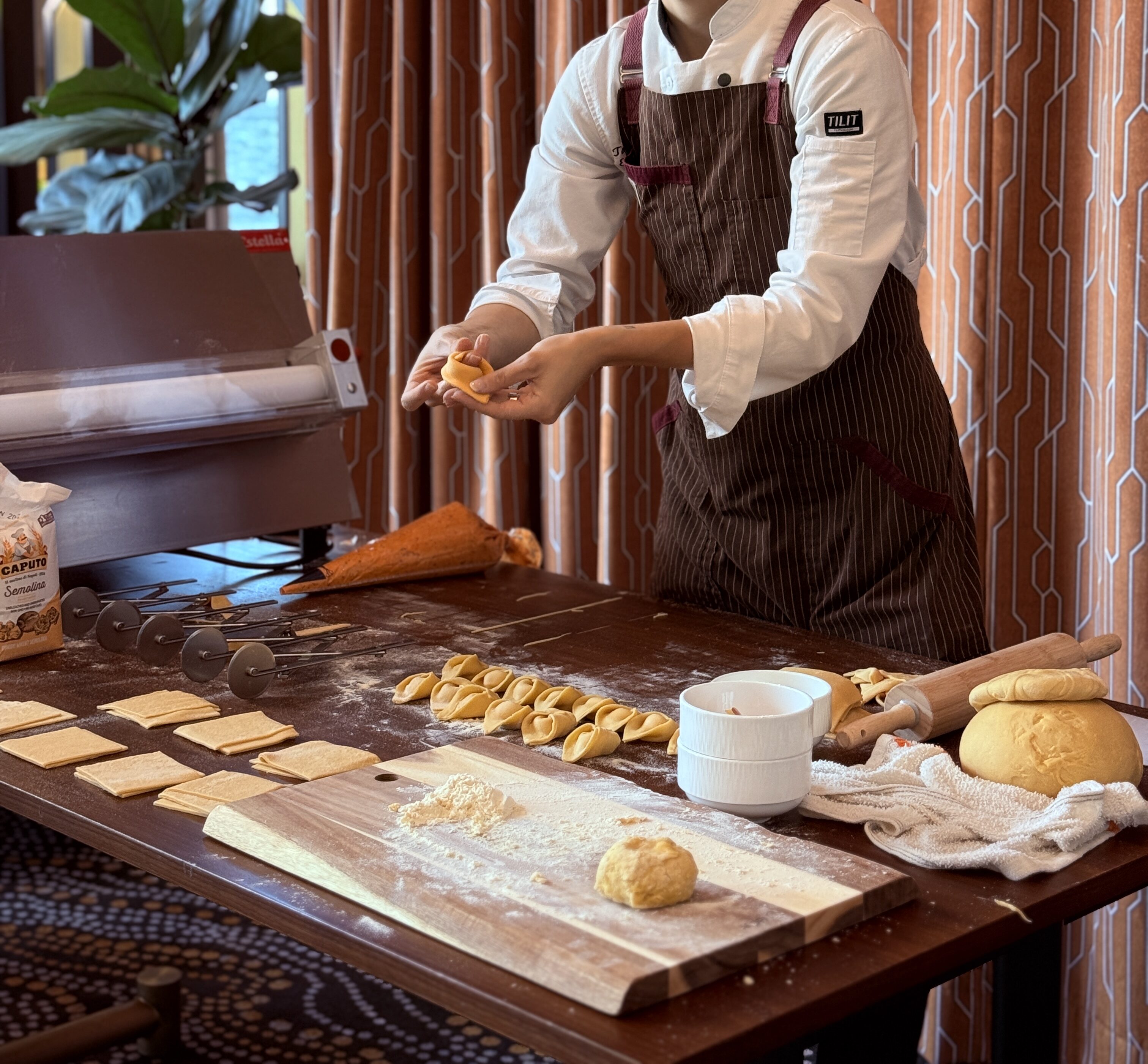 Chef Taylor, in a white coat and striped apron, prepares pasta by hand at a wooden table during a Social Club Pasta Making Class, surrounded by dough, pasta pieces, kitchen tools, and patterned curtains.