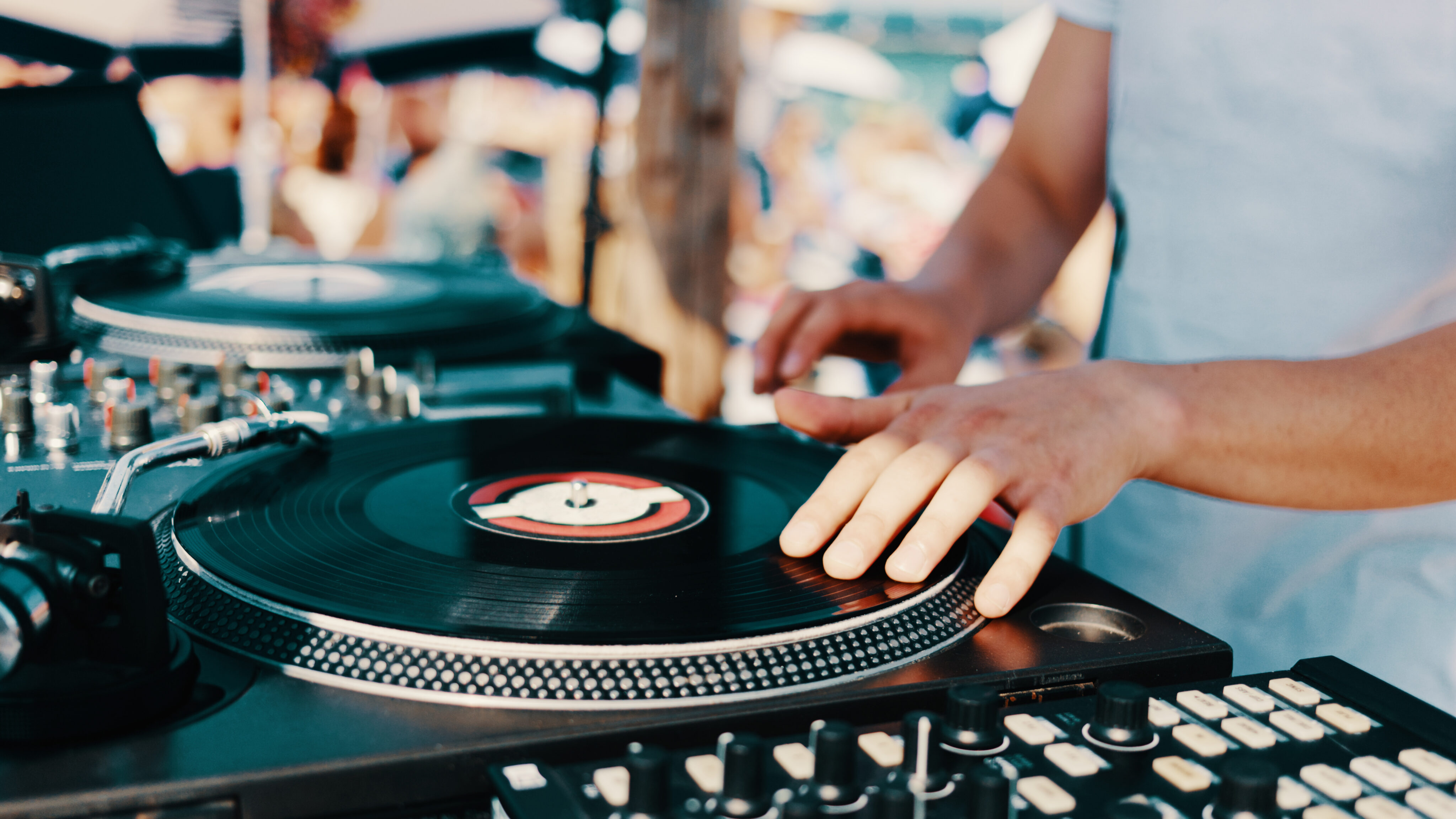 Close-up of a person's hands operating a turntable and mixing console, playing vinyl records at an outdoor event with a blurred crowd in the background.