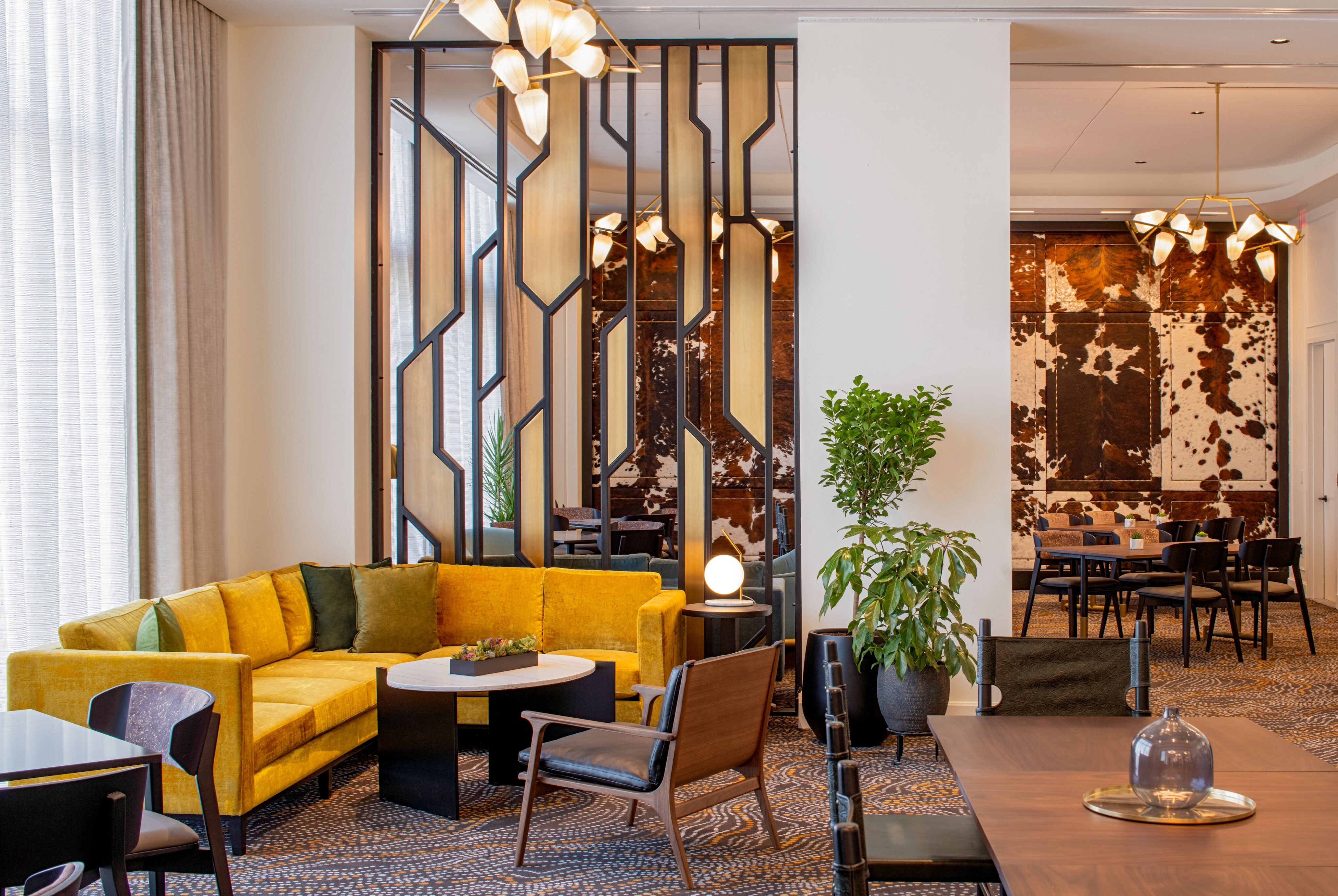 Modern lounge with a yellow sectional sofa, geometric partition, plants, and a dining area in the background with cowhide wall decor—perfect for hosting a Spanish Immersion experience or practicing the Spanish language in style.