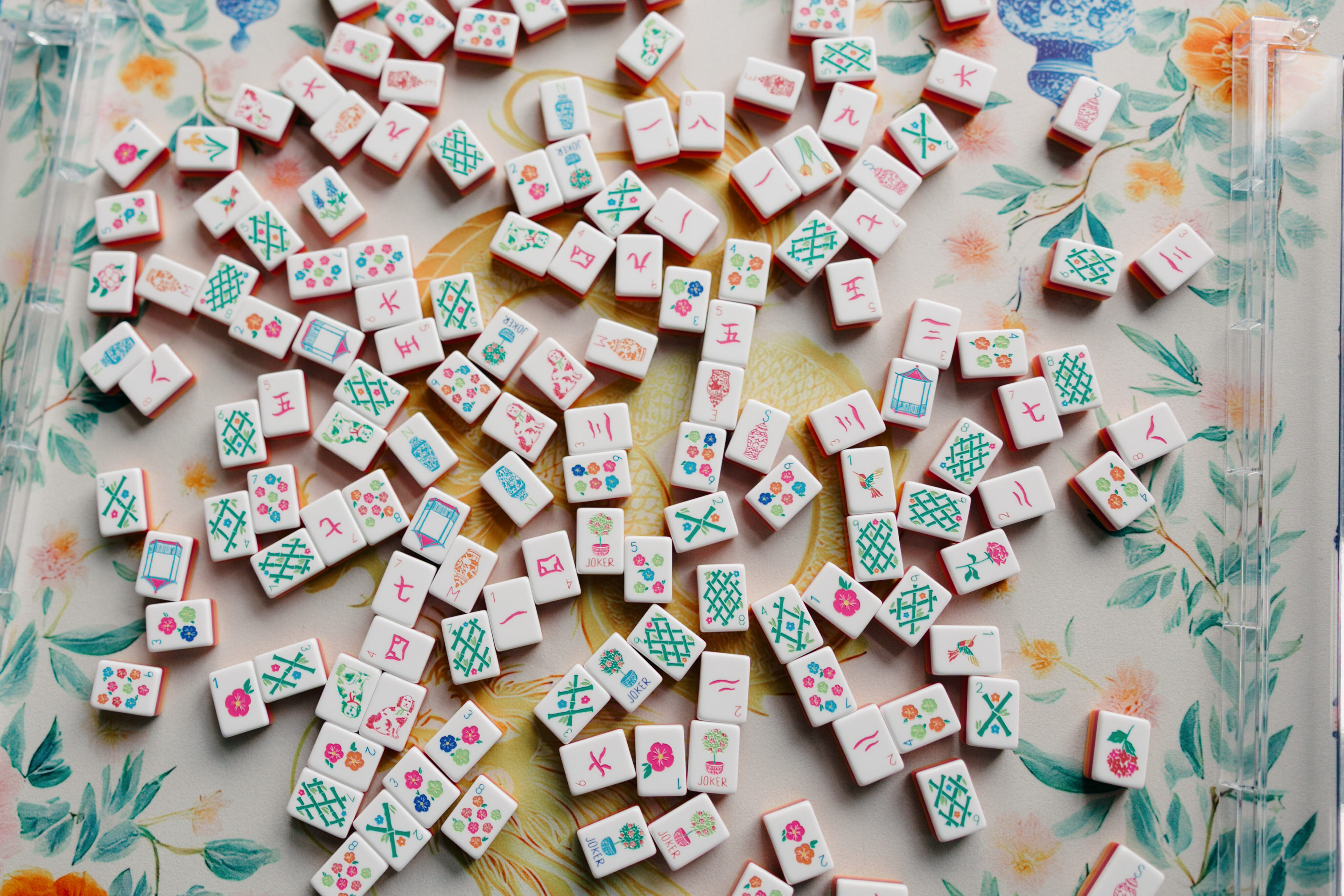 Scattered Mah Jongg tiles with various patterns and symbols rest on a floral background.